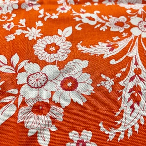 Könnte beinhalten: Orangener Stoff mit einem weißen Blumenmuster. Der Stoff hat eine strukturierte Oberfläche und ein sich wiederholendes Muster aus Blumen und Blättern.