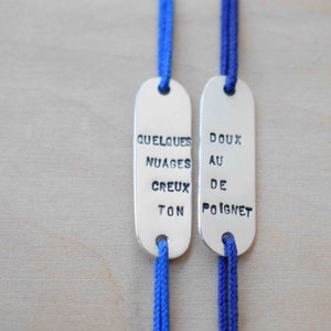 May include: Two silver charm bracelets with blue cords. The charms are oval shaped and have the text "Quelques Nuages Creux Ton" and "Doux Au De Poignet" engraved on them.