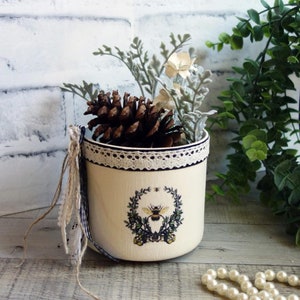 May include: A cream-colored decorative pot with a bee and floral design, filled with a pine cone and faux greenery. The pot has a lace trim and a blue and white ribbon. A string of pearls is in the foreground.