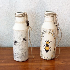 May include: Two white ceramic vases with a bee and floral design. The vases are decorated with twine and wooden beads.