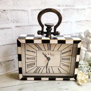 May include: A rectangular table clock with a black and white checkered border and a bronze-toned handle on top. The clock face features black Roman numerals and the text Nicole Miller Home. The clock hands are black and set against a white background.