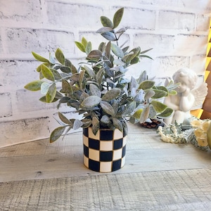 May include: A small artificial plant with sage green leaves in a cylindrical ceramic pot featuring a black and white checkered pattern. The plant has a frosted appearance on the foliage and is displayed in a vertical orientation with a flat base.