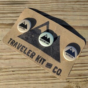 May include: Three round, cream-colored buttons with black mountain range designs and text. The buttons read "Explore", "Adventure", and "Wander". The buttons are on a brown card with the text "Traveler Kit Co."