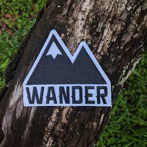 May include: Black and white embroidered patch featuring a mountain range design and the word "WANDER" in white lettering. The patch is attached to a tree trunk.