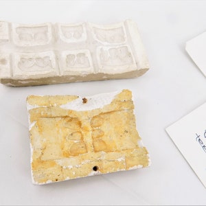May include: Two vintage plaster molds for making decorative items. One mold is white and has a rectangular shape with multiple squares containing a floral design. The other mold is yellow and has a rectangular shape with a design of three rounded shapes.
