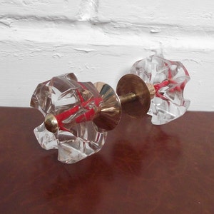 May include: A pair of clear glass door knobs with a faceted design and a brass connecting rod. The knobs have a red accent.