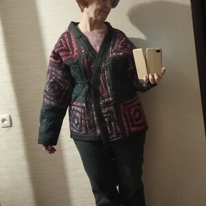 May include: A woman wearing a black and purple patterned jacket with a tie belt, black pants, and white sneakers. She is holding two cell phones in her hands.