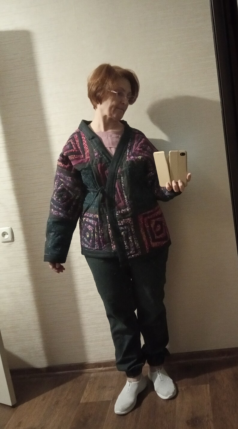 May include: A woman wearing a black and purple patterned jacket with a tie belt, black pants, and white sneakers. She is holding two cell phones in her hands.
