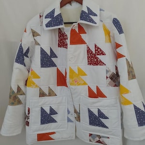 Könnte beinhalten: Eine weiße Steppjacke mit Patchwork-Design. Die Jacke hat einen Kragen, zwei Taschen und Knopfverschlüsse. Der Patchwork-Stoff enthält Dreiecke in blauen, orangefarbenen, gelben und roten Mustern. Die Jacke hängt an einem Holzbügel.