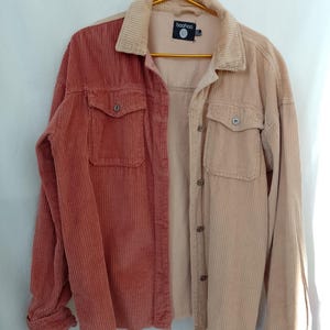 Peut inclure: Une chemise en velours côtelé bicolore, moitié rouge-brun, moitié beige clair. Elle comporte deux poches de poitrine, des boutons et l'étiquette de la marque "boohoo". Le tissu côtelé est à rayures verticales.