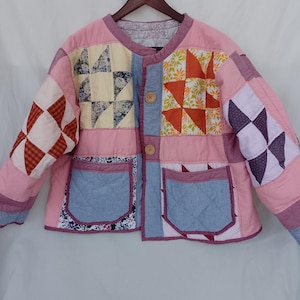 May include: A quilted jacket featuring a patchwork design with various floral and geometric patterns in shades of pink, orange, blue, and white. The jacket has a round neckline, long sleeves, two front pockets, and two light brown wooden buttons.