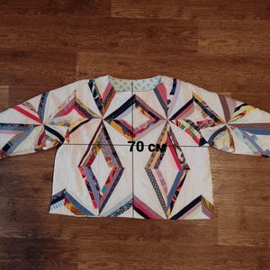 May include: A quilted jacket with a geometric design. The jacket is white with colorful fabric strips arranged in diamond and starburst patterns. The jacket measures 70 cm across.
