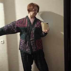 May include: A woman wearing a black and purple patterned kimono-style jacket with a black belt, black pants, and white shoes. She is holding a gold phone in her right hand.
