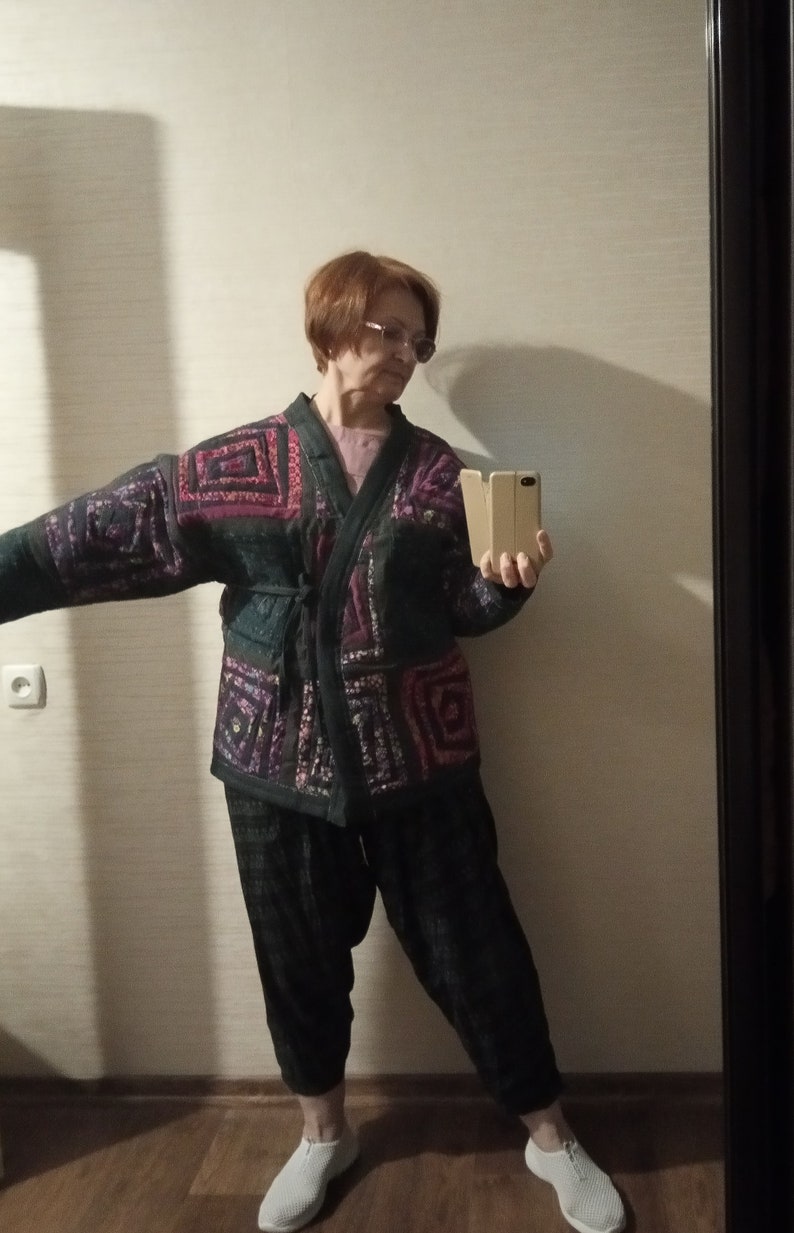 May include: A woman wearing a black and purple patterned kimono-style jacket with a black belt, black pants, and white shoes. She is holding a gold phone in her right hand.