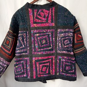 May include: A black quilted jacket with a patchwork design. The jacket is made of different fabrics with floral patterns in shades of pink, purple, and green. The jacket has a zipper closure and long sleeves.