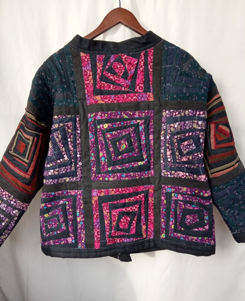 May include: A black quilted jacket with a patchwork design. The jacket is made of different fabrics with floral patterns in shades of pink, purple, and green. The jacket has a zipper closure and long sleeves.