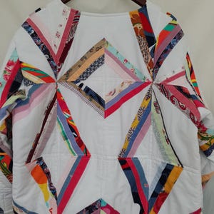 May include: White quilted jacket with colorful patchwork accents. The jacket features a geometric design with various fabric patterns in red, blue, yellow, and pink. It has long sleeves and two front pockets, hanging on a wooden hanger.