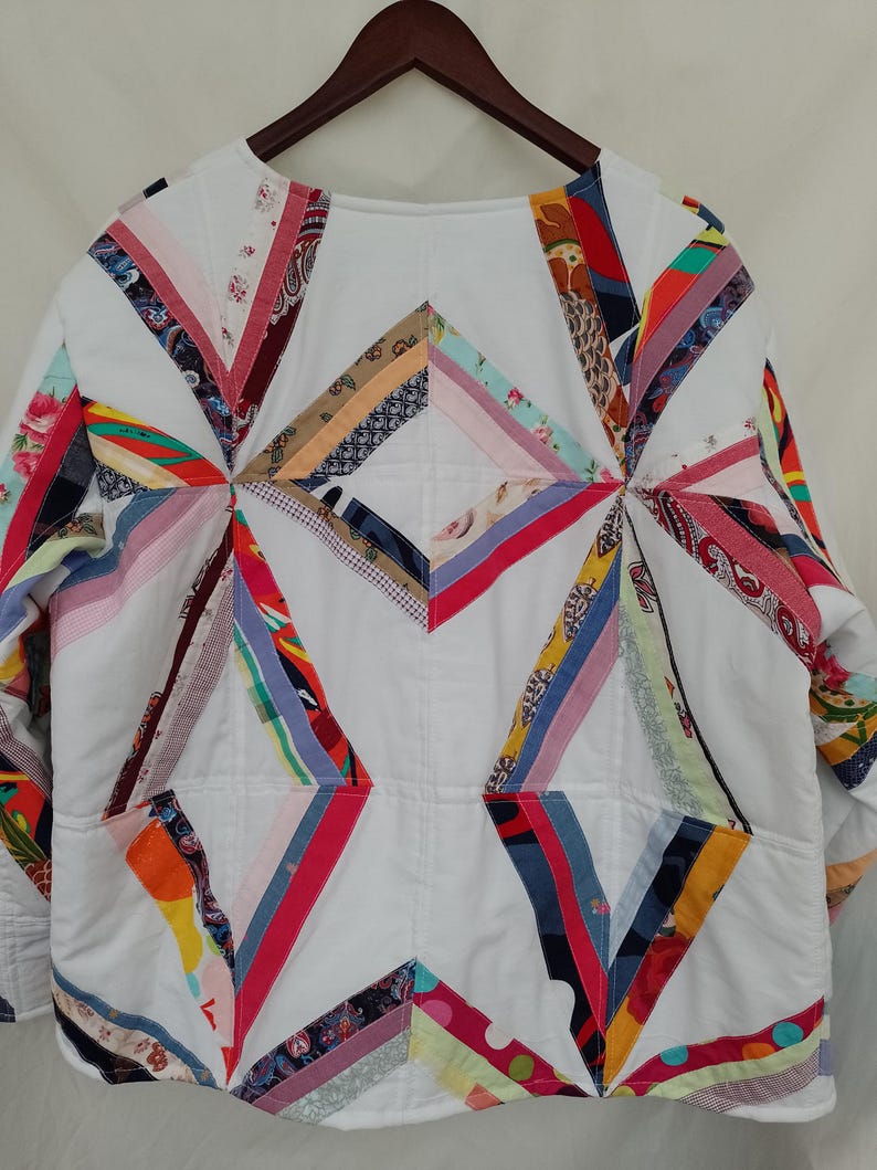 May include: White quilted jacket with colorful patchwork accents. The jacket features a geometric design with various fabric patterns in red, blue, yellow, and pink. It has long sleeves and two front pockets, hanging on a wooden hanger.