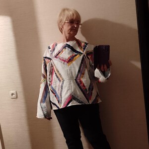 May include: A white patchwork top with geometric patterns in various colors, including red, blue, and yellow. The top is paired with black pants and white sneakers. The overall style is casual and handmade.