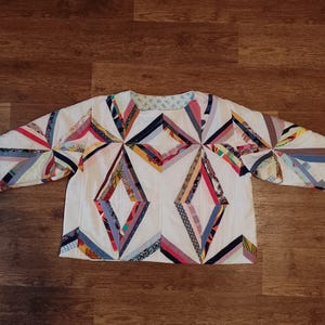 May include: A white, long-sleeved quilted jacket with a geometric diamond pattern. The design incorporates various colorful fabric strips in red, blue, yellow, and other hues. The jacket is laid flat on a wooden surface.