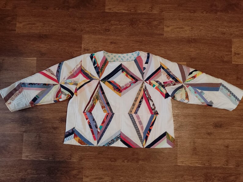 May include: A white, long-sleeved quilted jacket with a geometric diamond pattern. The design incorporates various colorful fabric strips in red, blue, yellow, and other hues. The jacket is laid flat on a wooden surface.