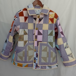 May include: A long-sleeve jacket constructed from various quilted fabric patches in shades of lavender, cream, orange, and floral patterns. The jacket features a collared neckline, a front button closure with four brown buttons, and two large front pockets.