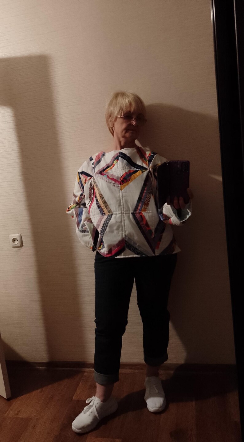 May include: A white quilted top with a geometric patchwork design in various colors, paired with dark blue jeans and white sneakers. The top features a unique, handcrafted aesthetic, showcasing a blend of textures and patterns.