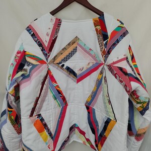 May include: White quilted jacket with colorful patchwork accents. The jacket features a geometric design with various fabric strips in shades of red, blue, yellow, and pink. The jacket is hanging on a wooden hanger.