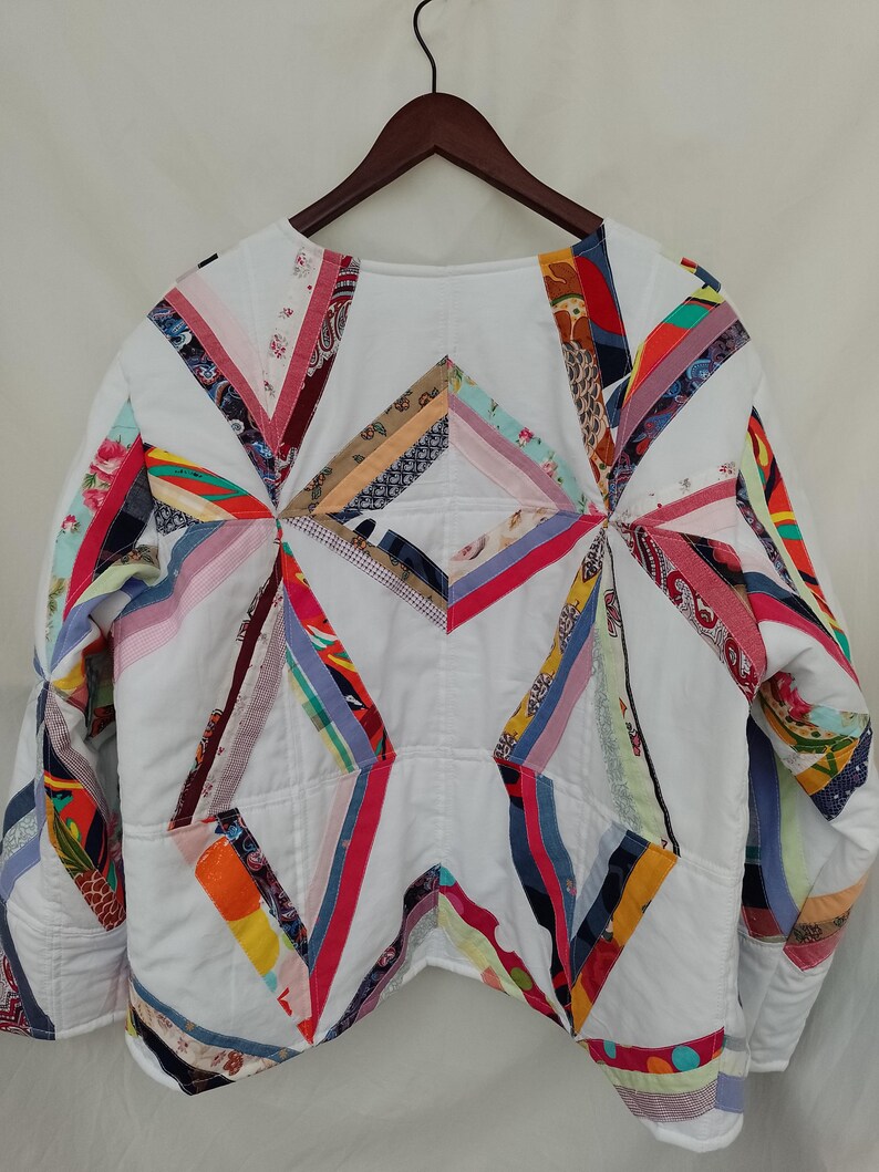 May include: White quilted jacket with colorful patchwork accents. The jacket features a geometric design with various fabric strips in shades of red, blue, yellow, and pink. The jacket is hanging on a wooden hanger.