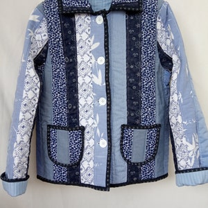 Puede incluir: Una chaqueta acolchada azul con ribete negro y botones blancos. La chaqueta tiene un diseño de patchwork con diferentes patrones de tela azul y blanca.