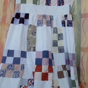 Peut inclure: Robe blanche sans manches avec un motif patchwork. La robe présente un corsage avec un décolleté à cordon de serrage et une jupe à volants. Le patchwork incorpore divers tissus à motifs dans les tons de bleu, vert, rose et marron.