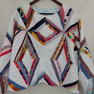 May include: A white quilted jacket with a geometric design. The jacket features colorful fabric strips in various patterns and colors, including red, blue, and yellow, arranged in a starburst pattern. The jacket has long sleeves and a round neckline.