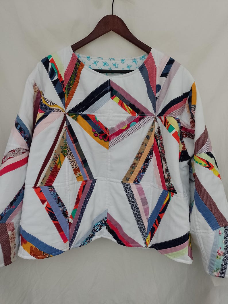 May include: A white quilted jacket with a geometric design. The jacket features colorful fabric strips in various patterns and colors, including red, blue, and yellow, arranged in a starburst pattern. The jacket has long sleeves and a round neckline.