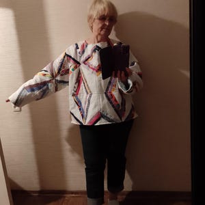 May include: A quilted jacket with a geometric pattern in various colors, including red, blue, and yellow, is paired with dark blue jeans and white sneakers. The jacket's design features a patchwork style.