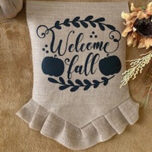 Könnte beinhalten: Ein Willkommen Herbst-Banner aus Burlap mit schwarzer Schrift und zwei schwarzen Kürbis-Silhouetten. Das Banner hat einen gekräuselten Rand.