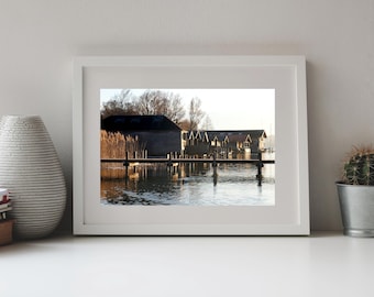 May include: A white framed landscape photograph depicting a wooden pier extending over calm water toward dark boathouses at sunset. Tall reeds grow along the shoreline near the structures, and the scene is illuminated by warm, golden light.