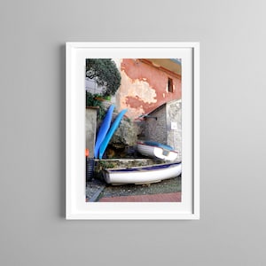 May include: A framed photograph of a coastal scene featuring several small boats resting on the ground. The boats are white with blue accents. Tall, bright blue surfboards lean against a stone wall. The background includes a weathered building with a red facade.