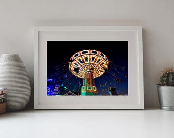 May include: A white framed wall art print featuring a night scene of a brightly illuminated carnival swing ride. The ride displays vibrant orange, yellow, and blue lights against a dark sky, with suspended seats rotating around a central tower.