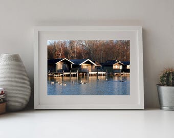 May include: A framed landscape photograph featuring a row of dark wooden boathouses on stilts over a calm blue lake, with several ducks swimming in the foreground and a dense forest of bare trees in the background under golden sunlight.