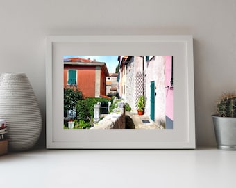 May include: A framed photograph of a narrow alleyway between buildings with orange and pink stucco walls, green shutters, a stone wall, and a potted plant. The scene is captured in bright daylight, showcasing the architectural details of the structures.