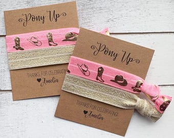 May include: Two brown cards with the text "Pony Up" and "Thanks for Celebrating! Amelia". Each card has a pink elastic hair tie with cowboy boot, hat, and horseshoe designs. A gold elastic band is also included.