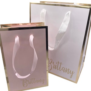 K&ouml;nnte beinhalten: Zwei Geschenkt&uuml;ten mit goldenen R&auml;ndern und wei&szlig;en Schleifenb&auml;ndern. Die gr&ouml;&szlig;ere Tasche ist wei&szlig; mit dem Namen "Brittany" in goldenen Buchstaben. Die kleinere Tasche ist rosa mit dem Namen "Brittany" in goldenen Buchstaben.