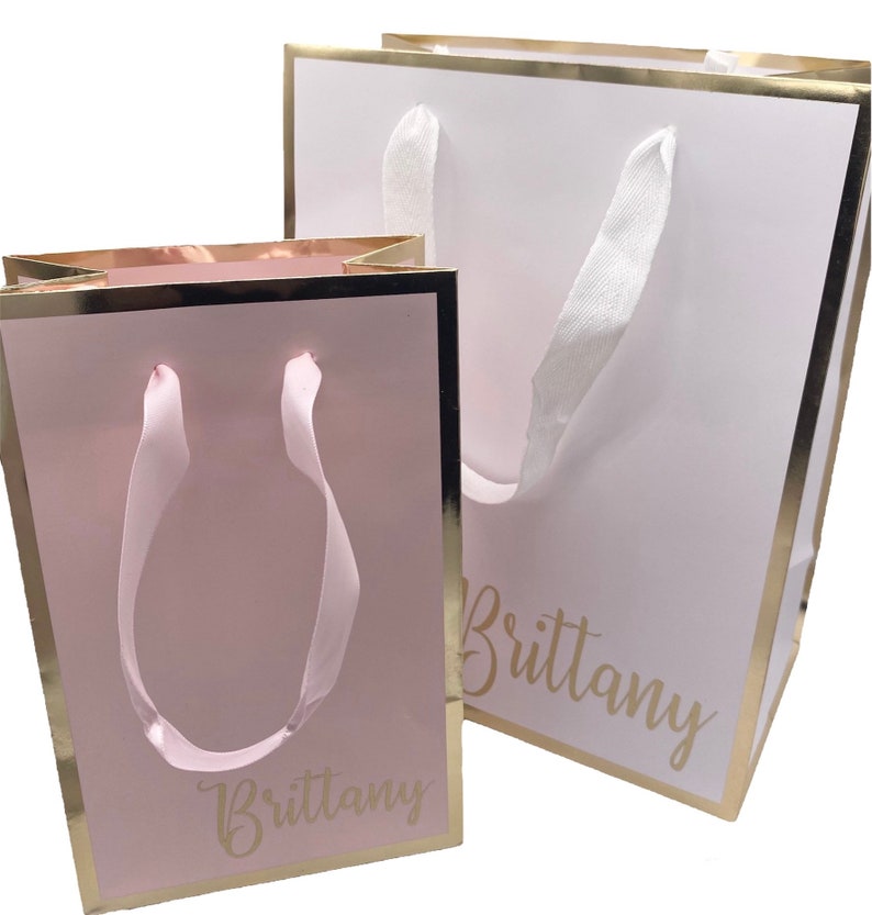K&ouml;nnte beinhalten: Zwei Geschenkt&uuml;ten mit goldenen R&auml;ndern und wei&szlig;en Schleifenb&auml;ndern. Die gr&ouml;&szlig;ere Tasche ist wei&szlig; mit dem Namen "Brittany" in goldenen Buchstaben. Die kleinere Tasche ist rosa mit dem Namen "Brittany" in goldenen Buchstaben.