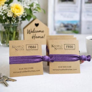 May include: Several business card holders with purple elastic bands. Each card is tan with the Kelsie Stites and Real of Ohio logos, a phone number, and an email address. A wooden house-shaped sign with the words "Welcome Home!" is in the background.