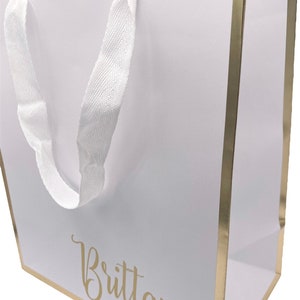 K&ouml;nnte beinhalten: Wei&szlig;e Geschenkt&uuml;te mit goldfarbenem Rand und wei&szlig;en Schleifenb&auml;ndern. Die Tasche tr&auml;gt den Namen "Brittany" in goldenen Lettern.
