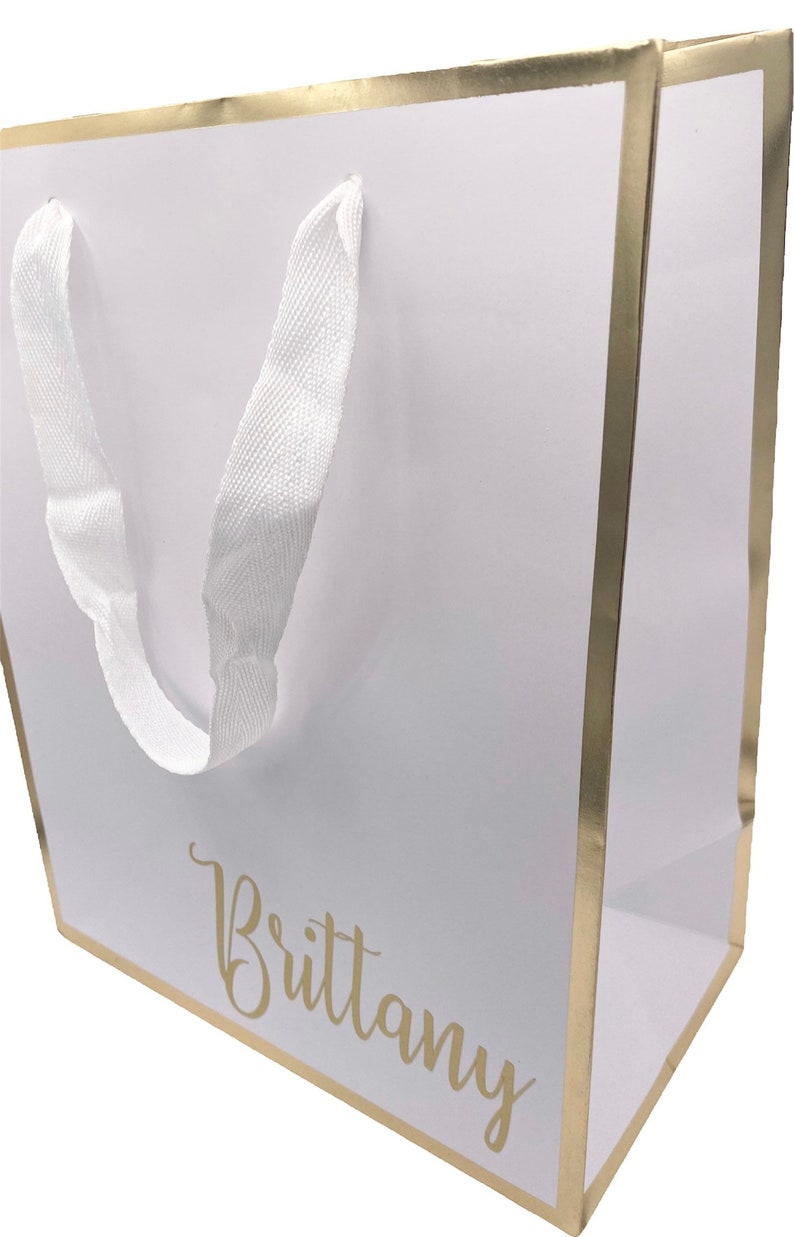 K&ouml;nnte beinhalten: Wei&szlig;e Geschenkt&uuml;te mit goldfarbenem Rand und wei&szlig;en Schleifenb&auml;ndern. Die Tasche tr&auml;gt den Namen "Brittany" in goldenen Lettern.