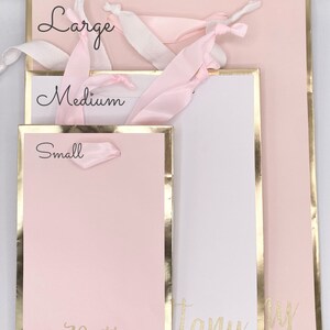 K&ouml;nnte beinhalten: Drei rosa Geschenkt&uuml;ten mit goldenen Verzierungen und wei&szlig;em Band. Die Taschen sind mit "Gro&szlig;", "Mittel" und "Klein" beschriftet. Die gr&ouml;&szlig;te Tasche tr&auml;gt den Namen "Brittany" in goldenen Glitzer auf der Vorderseite.