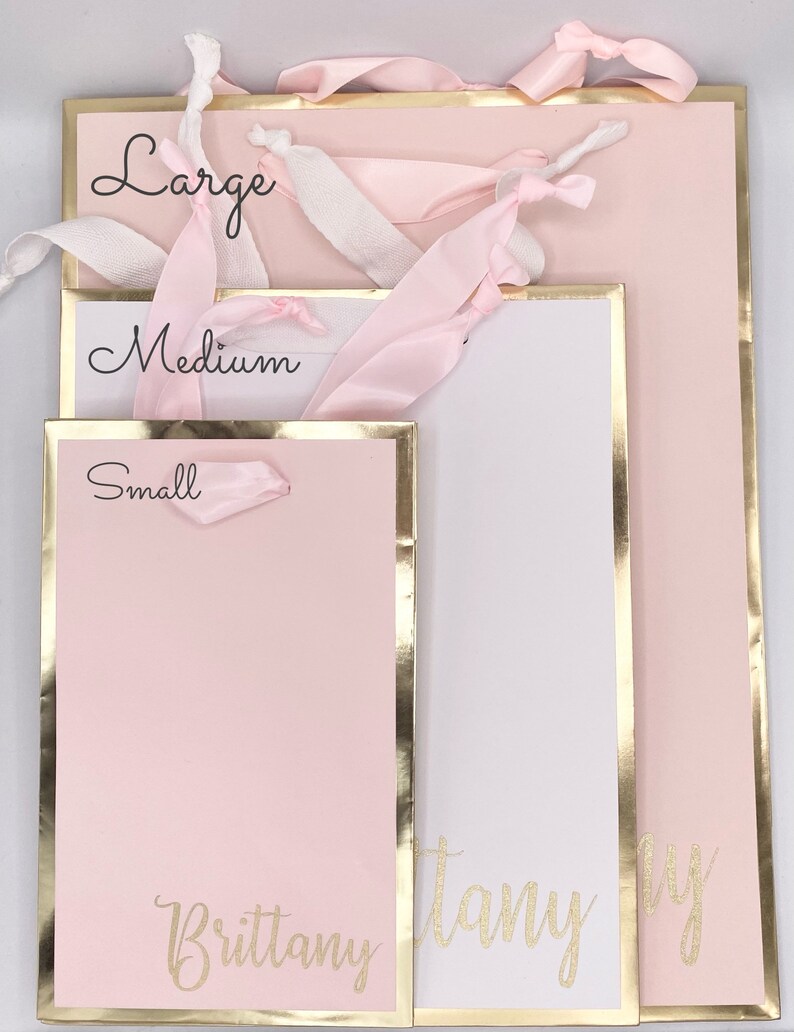 K&ouml;nnte beinhalten: Drei rosa Geschenkt&uuml;ten mit goldenen Verzierungen und wei&szlig;em Band. Die Taschen sind mit "Gro&szlig;", "Mittel" und "Klein" beschriftet. Die gr&ouml;&szlig;te Tasche tr&auml;gt den Namen "Brittany" in goldenen Glitzer auf der Vorderseite.