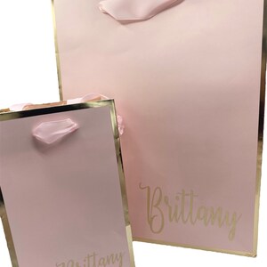 K&ouml;nnte beinhalten: Zwei rosa Geschenkt&uuml;ten mit goldenen R&auml;ndern und rosa Schleifenb&auml;ndern. Die Taschen sind mit dem Namen "Brittany" in goldenen Buchstaben personalisiert.