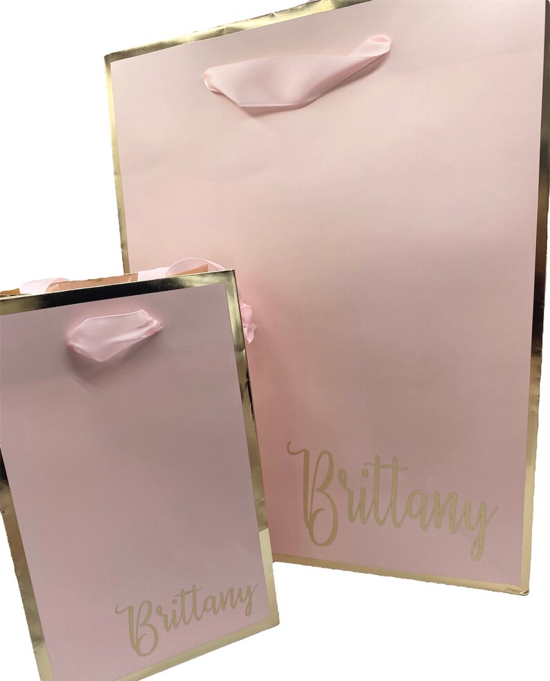K&ouml;nnte beinhalten: Zwei rosa Geschenkt&uuml;ten mit goldenen R&auml;ndern und rosa Schleifenb&auml;ndern. Die Taschen sind mit dem Namen "Brittany" in goldenen Buchstaben personalisiert.